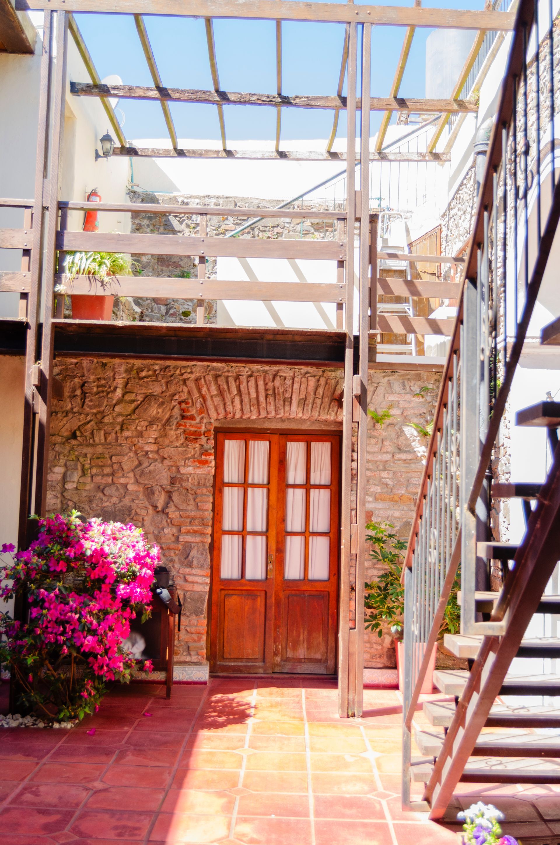 Un patio con un edificio de piedra que cuenta con puertas dobles de madera, suelos de baldosas rojas, una celosía de madera y una escalera metálica.