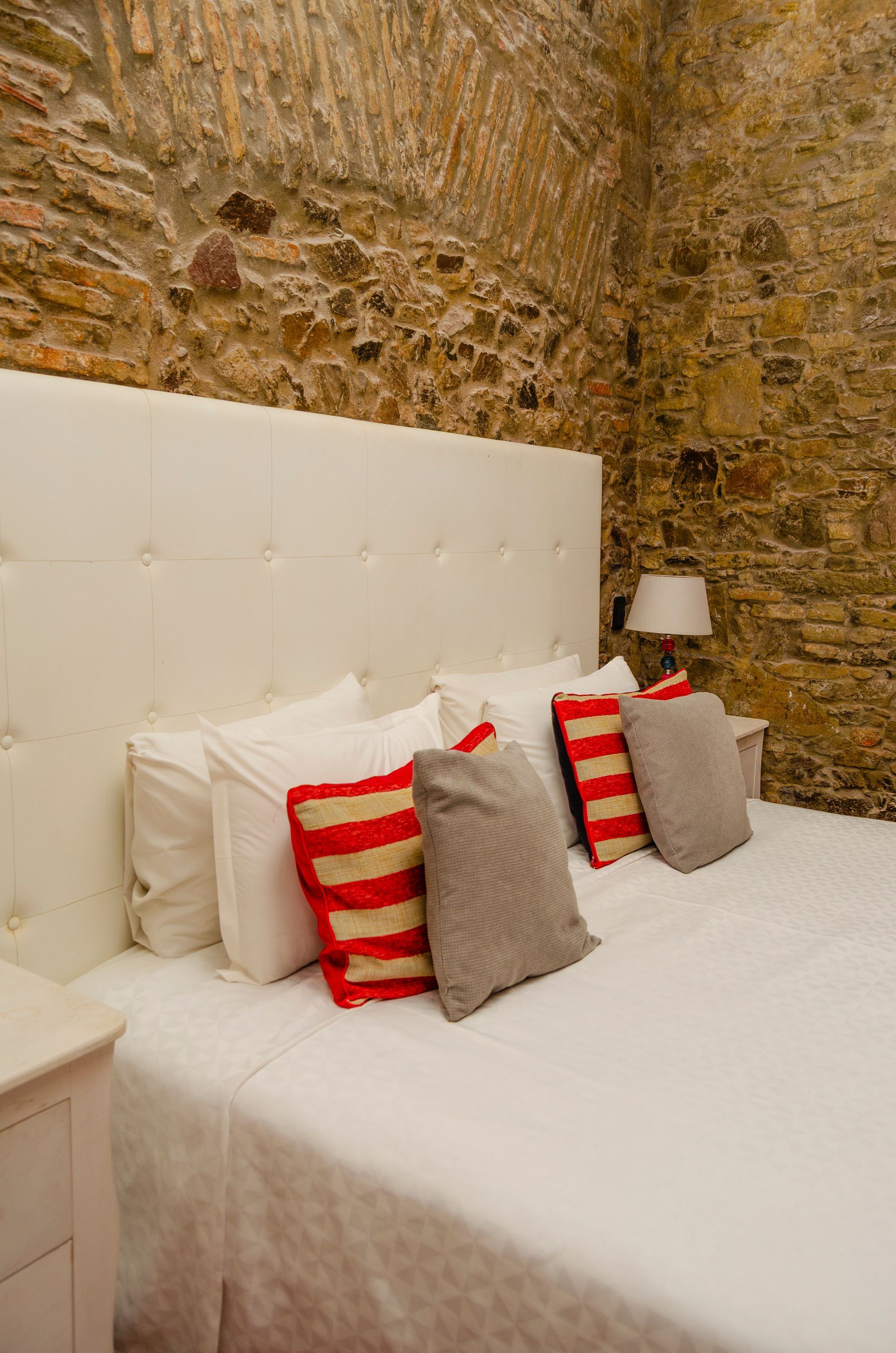 Una cama con sábanas blancas y cabecero acolchado, situada contra una pared de piedra rústica con una pequeña lámpara en la mesita de noche.