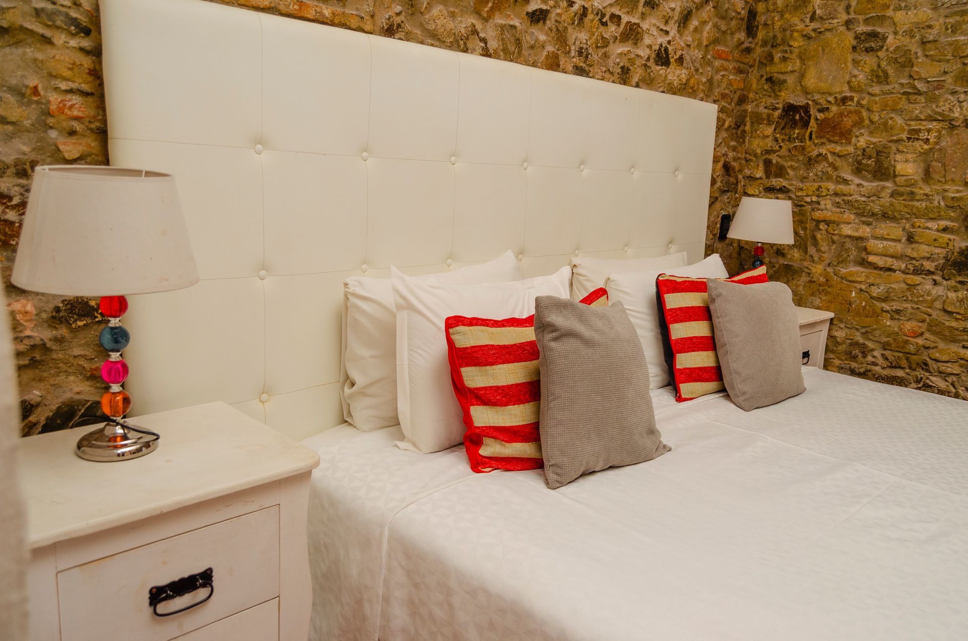 Un dormitorio con cabecero blanco acolchado, ropa de cama blanca y paredes de piedra. Junto a la cama hay una lámpara con cuentas de colores.