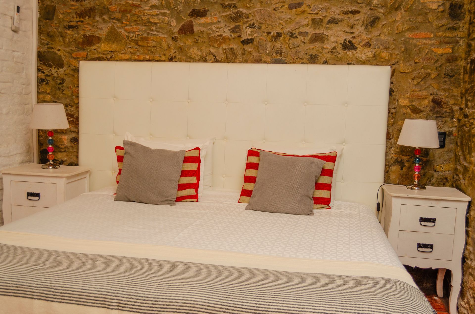 Una cama con ropa de cama blanca, almohadas a rayas grises y rojas, y un gran cabecero blanco contra una pared de piedra rústica.
