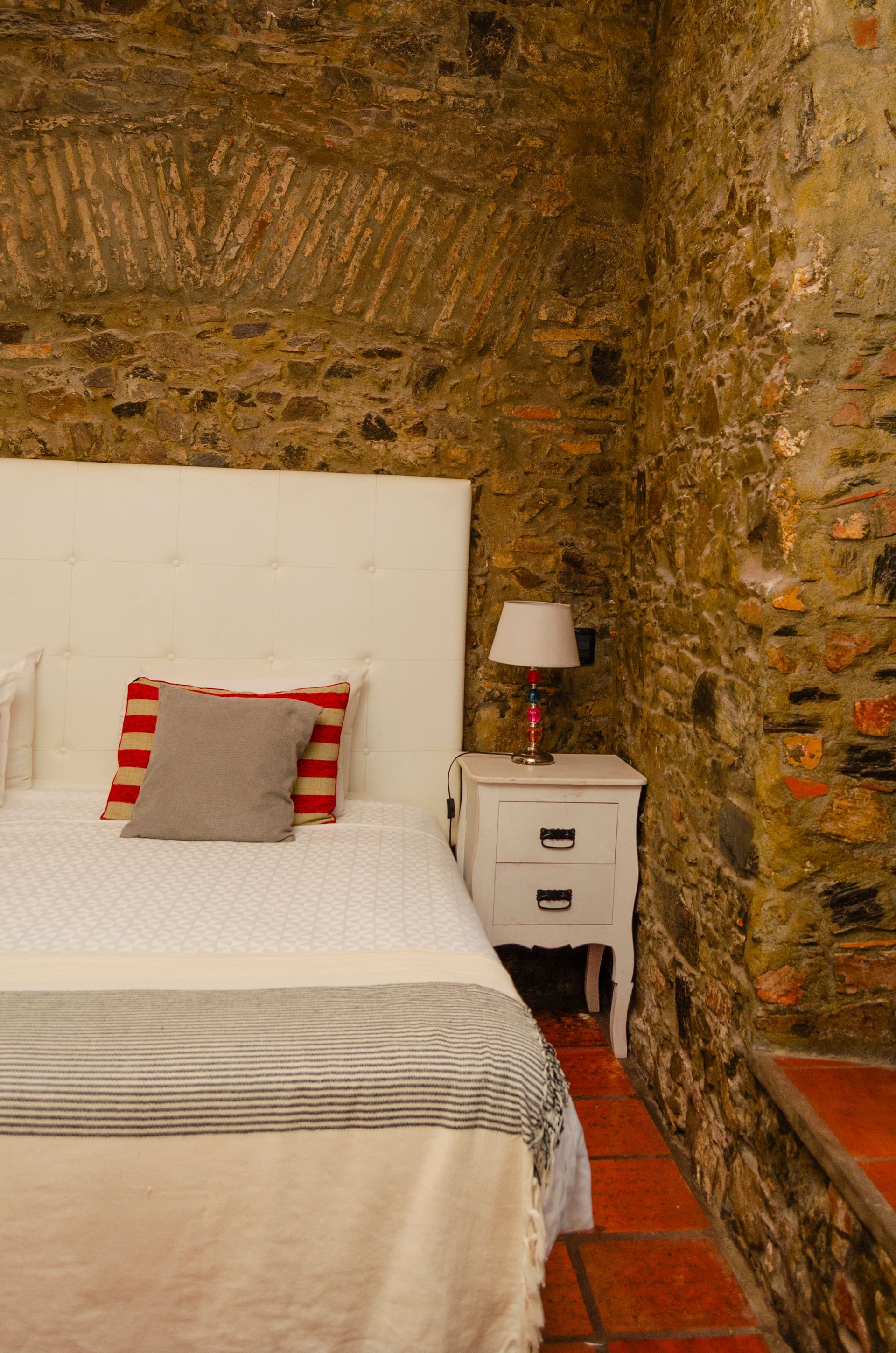 Una cama con cabecero blanco y almohadas a rayas se encuentra junto a una mesita de noche blanca, contra una pared rústica de piedra vista.