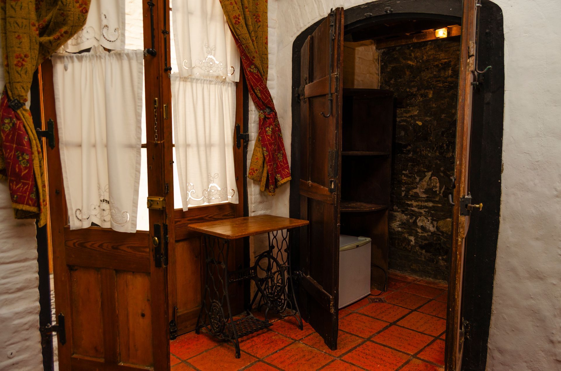 Una habitación con suelo de baldosas de terracota, una mesa con una máquina de coser antigua cerca de una puerta con cortinas y una alcoba con paredes de piedra.