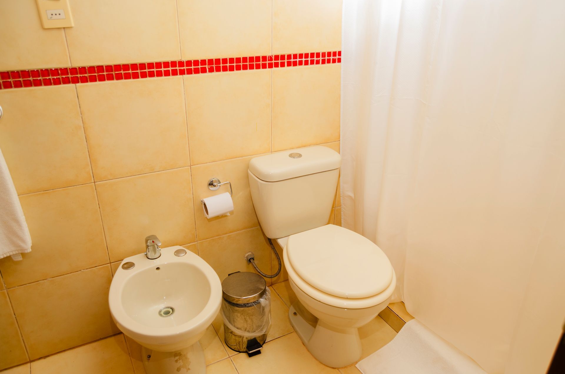 Un inodoro y un bidé blancos en un baño con paredes alicatadas en color beige y una franja decorativa roja horizontal.