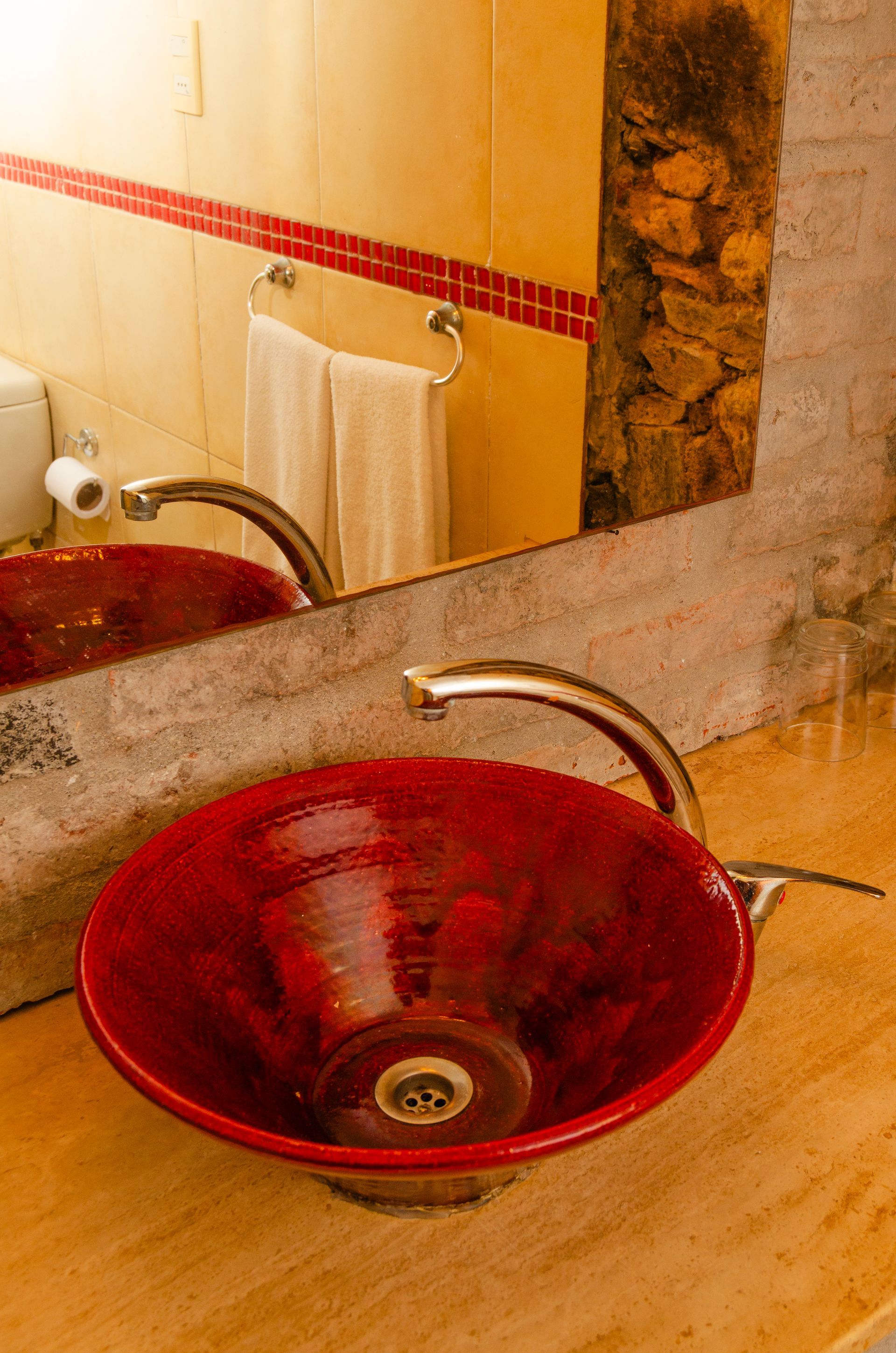 Un lavabo de color rojo brillante descansa sobre una encimera de piedra beige en un baño con detalles de ladrillo rústico y un espejo montado en la pared.