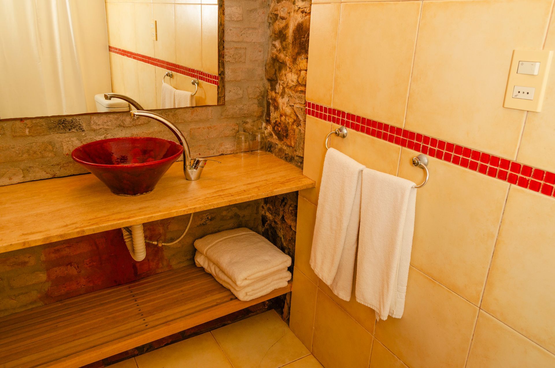 Un tocador de baño rústico con un lavabo de cristal rojo, encimeras de madera y toallas blancas colgadas en la pared alicatada.