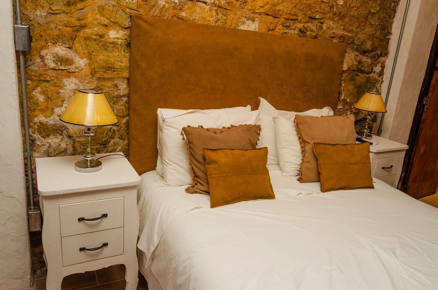Una cama blanca con almohadas marrones se apoya contra una pared de piedra rústica, flanqueada por dos mesitas de noche blancas con lámparas.