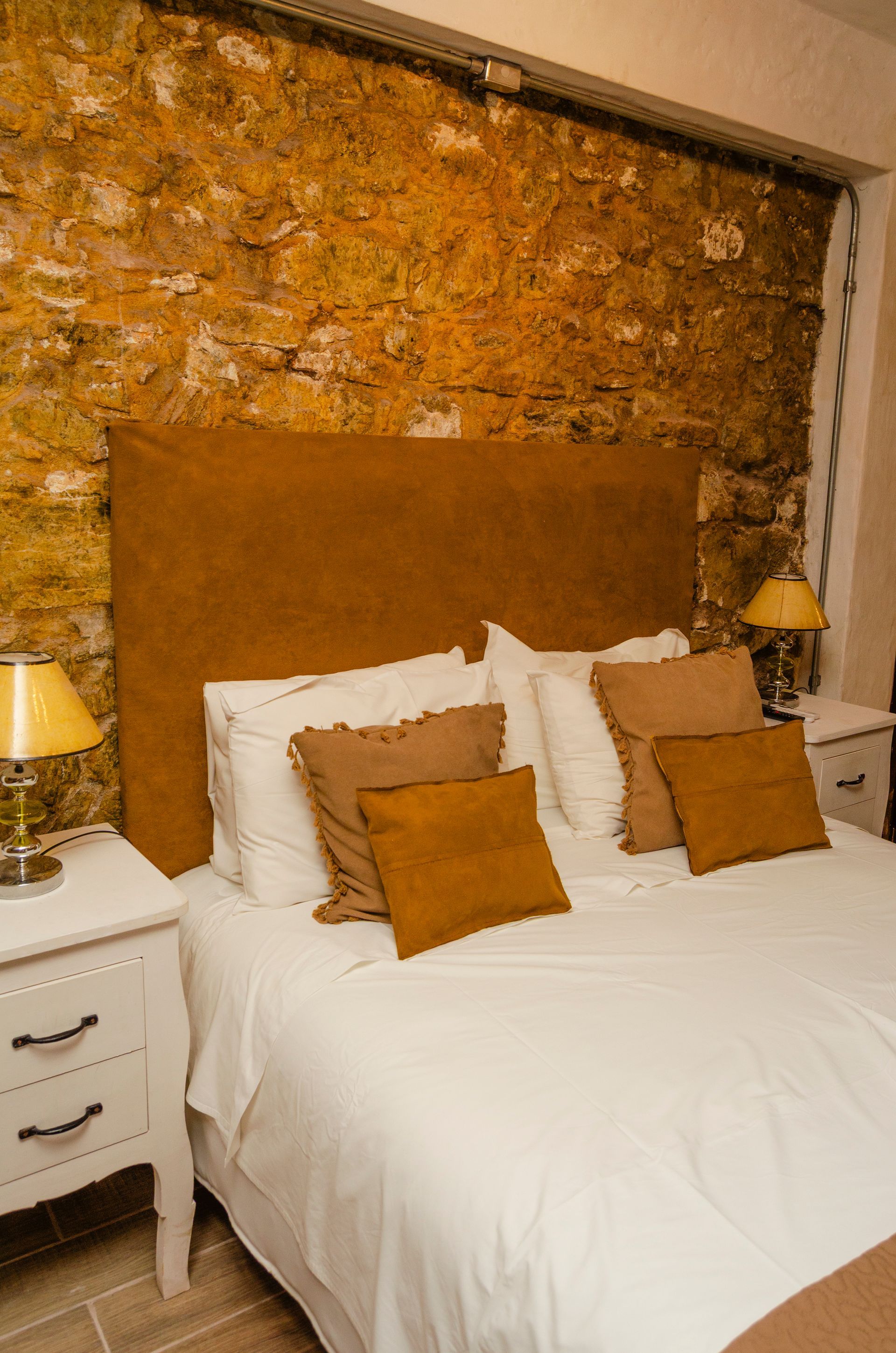 Una cama con cabecero dorado contra una pared de piedra rústica, flanqueada por mesitas de noche blancas con lámparas amarillas.