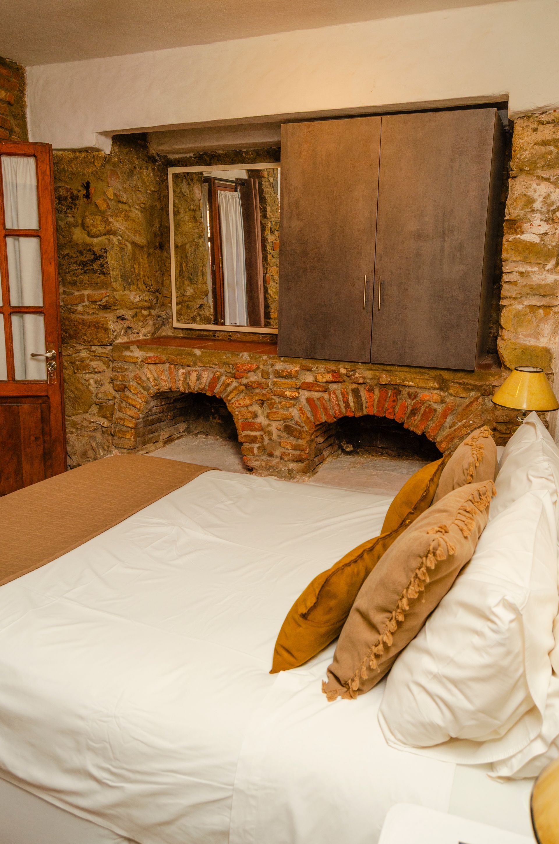 Un dormitorio rústico con una cama con sábanas blancas, cojines decorativos, una pared de piedra con dos arcos y un gran armario de madera.