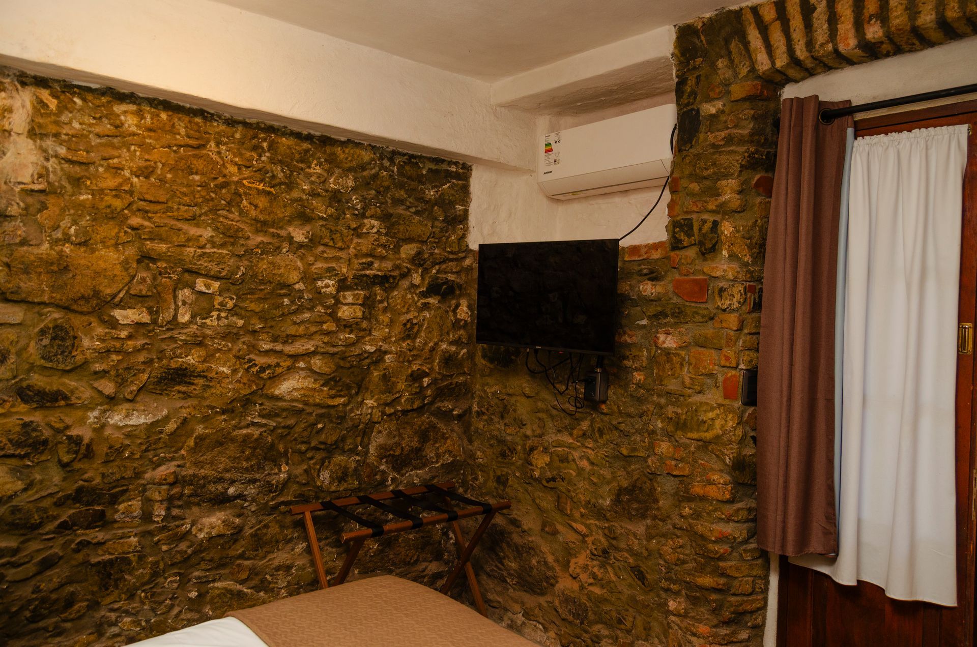 Un dormitorio con paredes de piedra rústica, un televisor montado en la pared, un pequeño perchero y una ventana con cortinas.