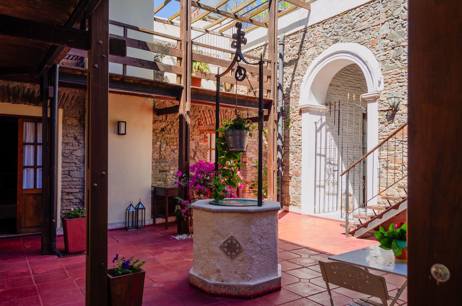 Un patio exterior de piedra cuenta con un pozo de los deseos en el centro, plantas en macetas y un suelo de baldosas rojas bajo una pérgola.