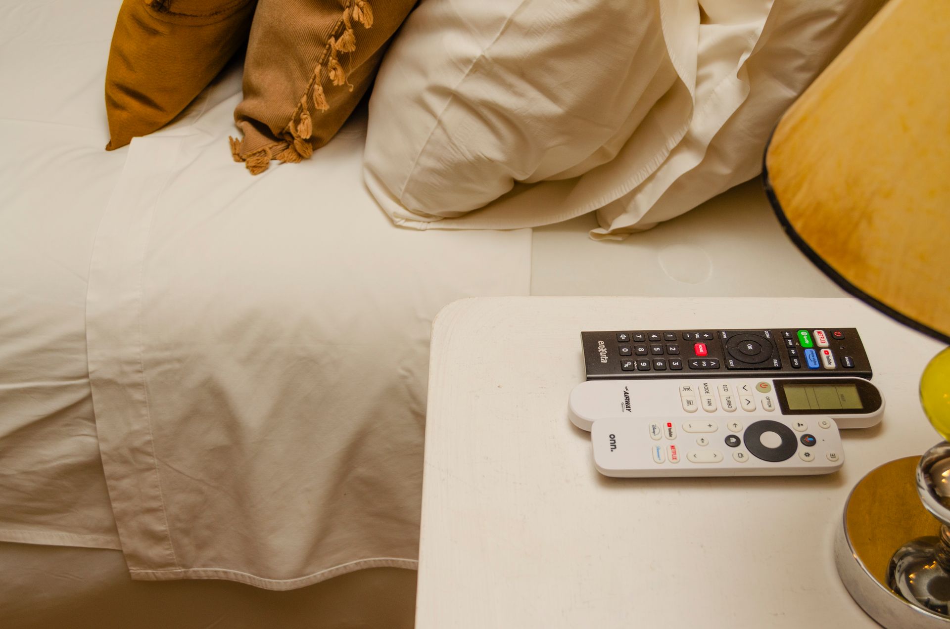 Tres mandos a distancia reposan sobre una mesita de noche blanca junto a una cama con sábanas blancas y almohadas color canela.