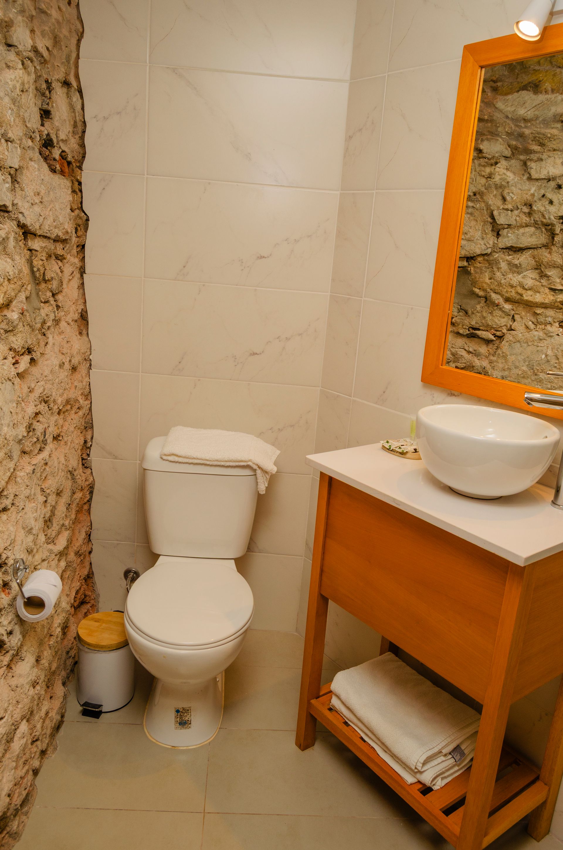 Un baño moderno cuenta con un inodoro blanco, un tocador de madera con lavabo sobre encimera y una pared rústica de piedra natural.