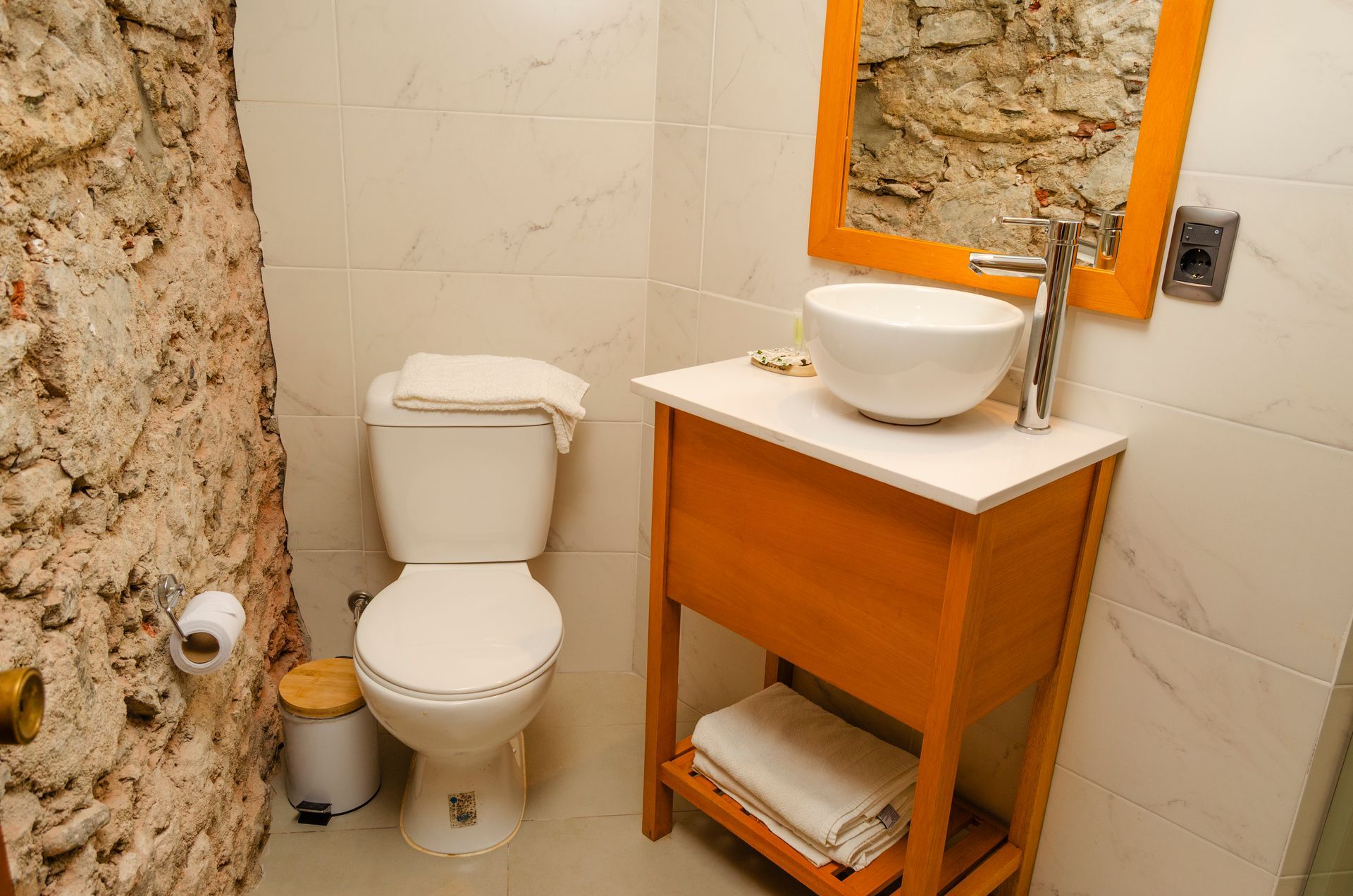 Un pequeño baño con inodoro blanco, tocador de madera con lavabo sobre encimera y pared de piedra rústica.
