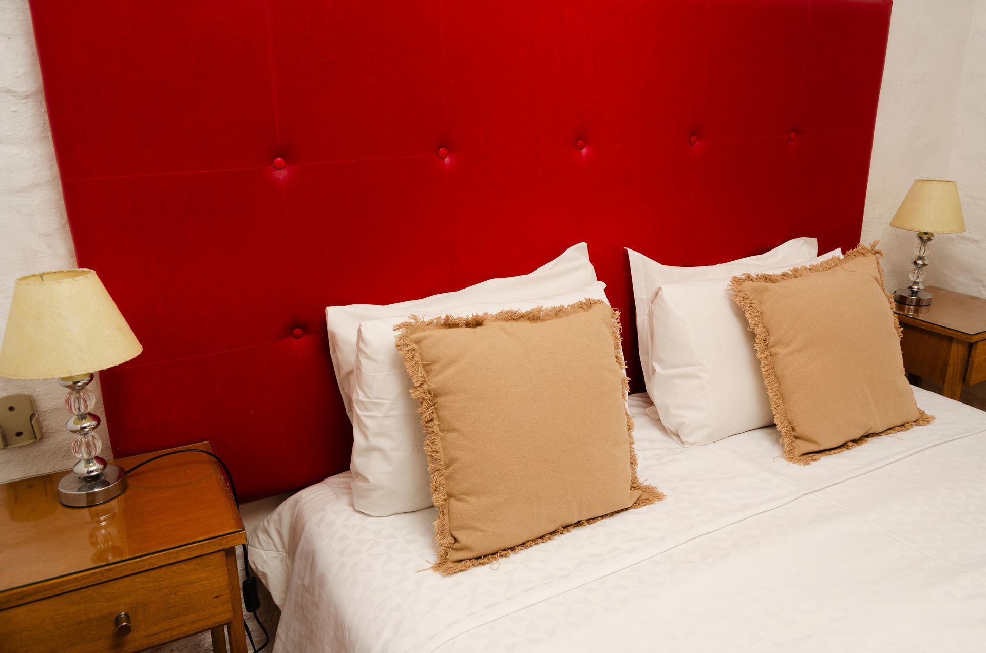 Una cama impecablemente hecha con sábanas blancas, cojines decorativos de color beige, un gran cabecero capitoné rojo y lámparas de noche de madera.