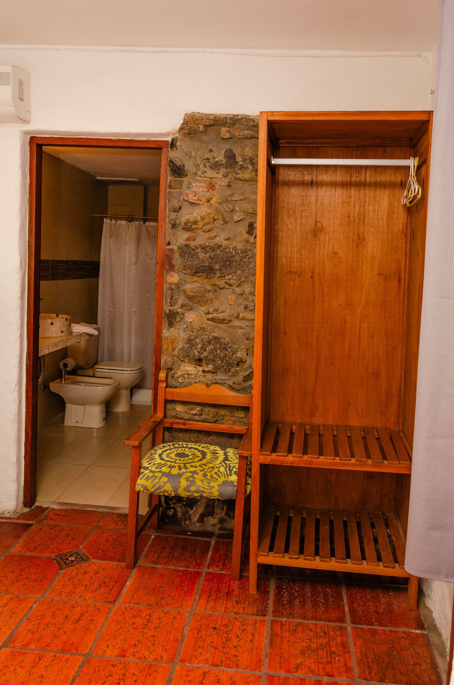 Un armario de madera abierto con estantes se encuentra junto a una silla y una puerta que da a un baño con inodoro y bidé.
