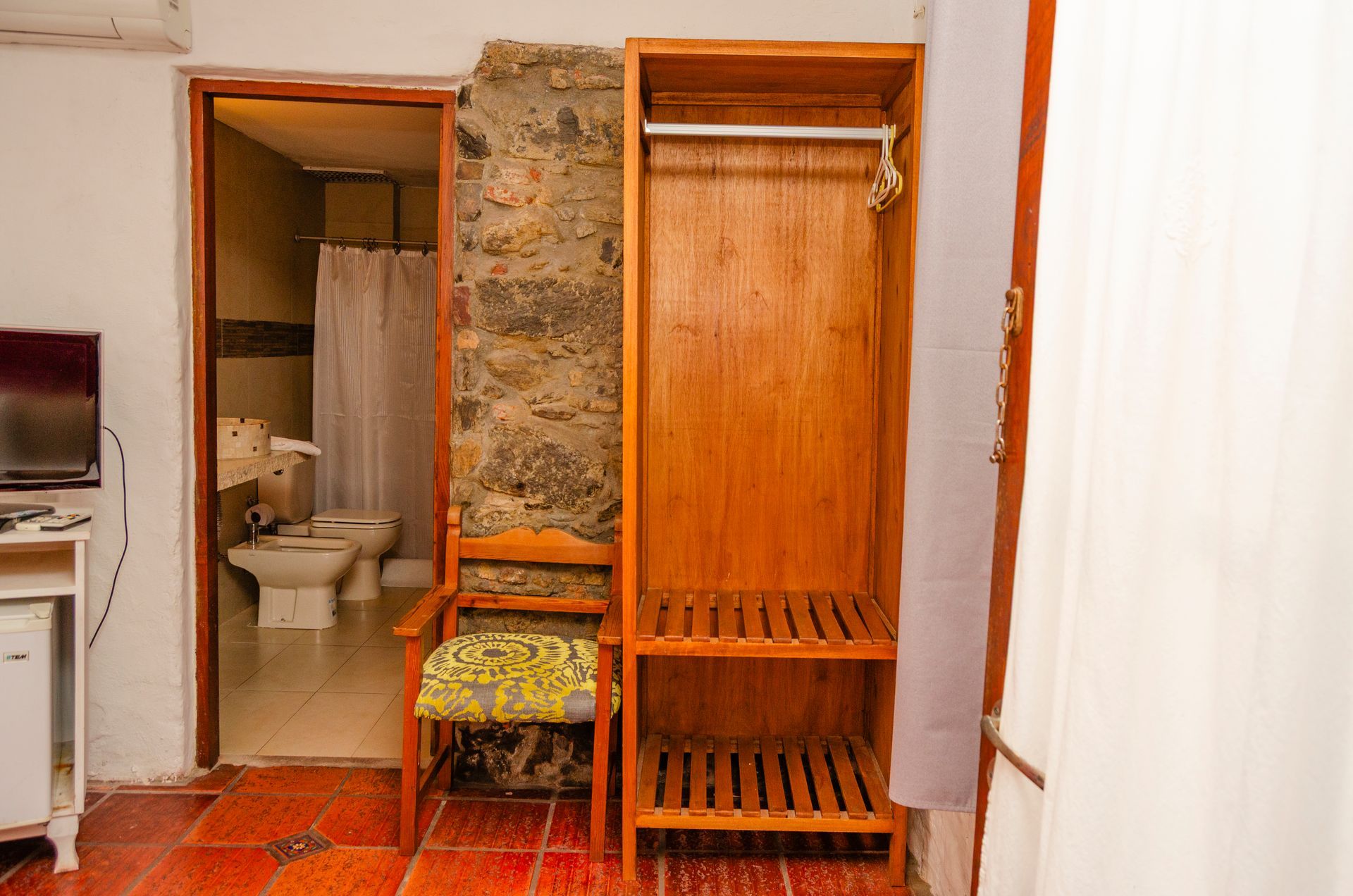 Un armario de madera abierto se encuentra junto a una puerta de piedra, dejando ver un baño con sanitarios blancos y suelo de baldosas.