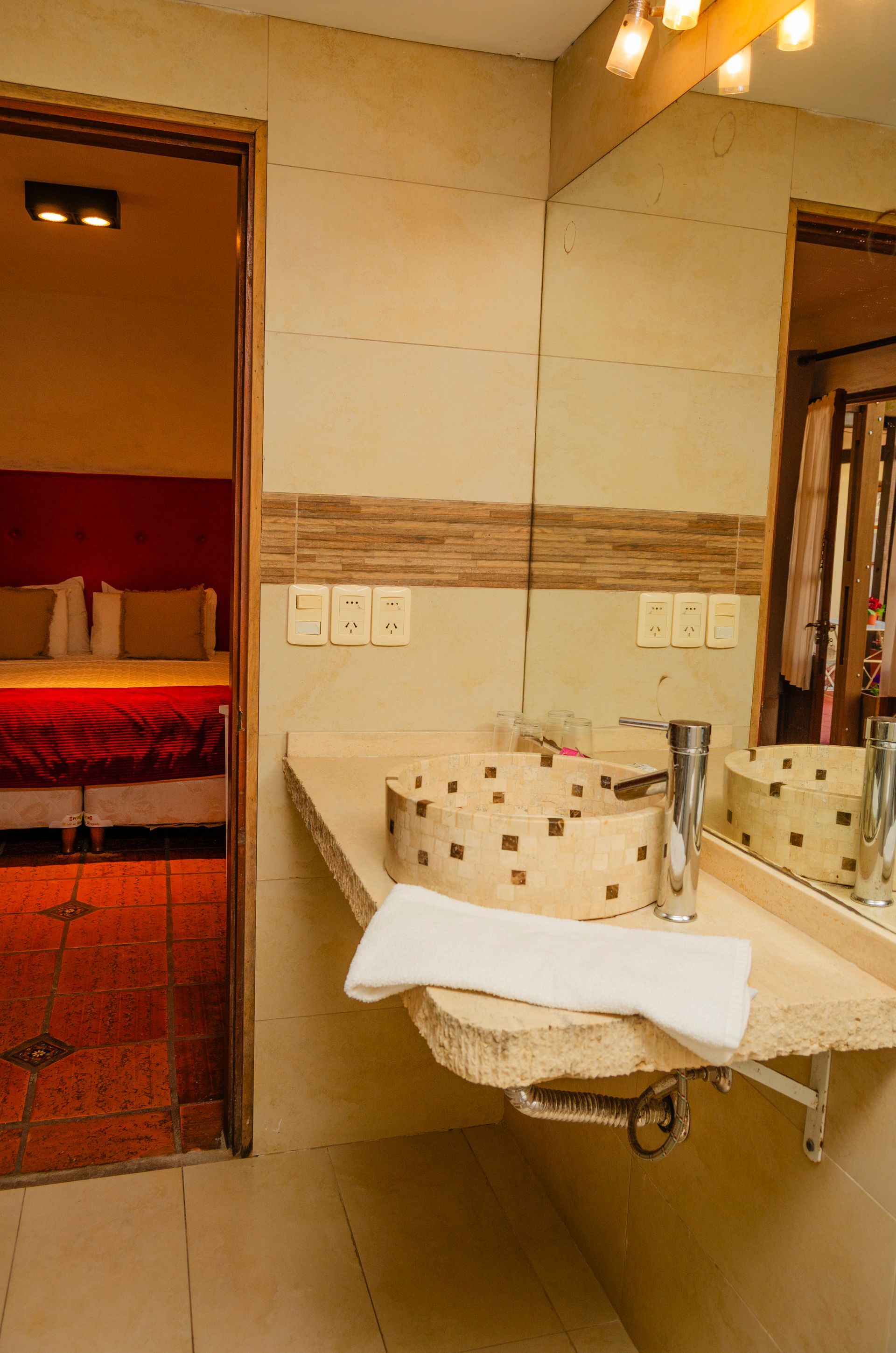 Un tocador de baño con lavabo sobre azulejos y una toalla blanca, que da a través de una puerta a un dormitorio con una cama roja.