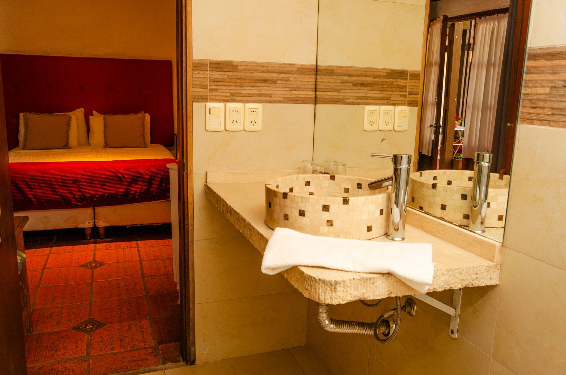 Un lavabo de piedra sobre una encimera en un baño con vistas a un dormitorio con una colcha roja y almohadas beige.
