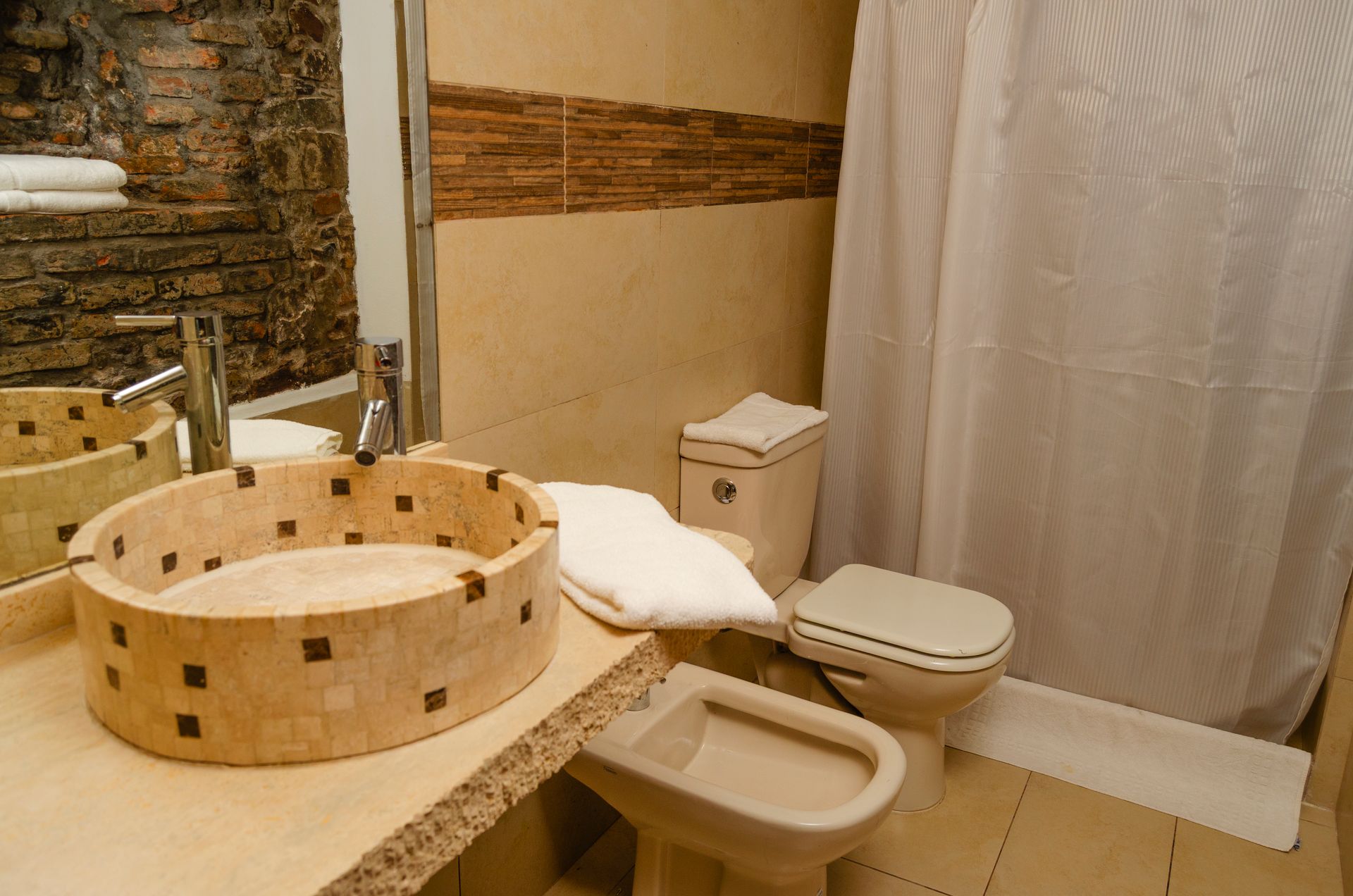 Un baño con lavabo circular de piedra sobre encimera, inodoro, bidé y ducha con cortina blanca.