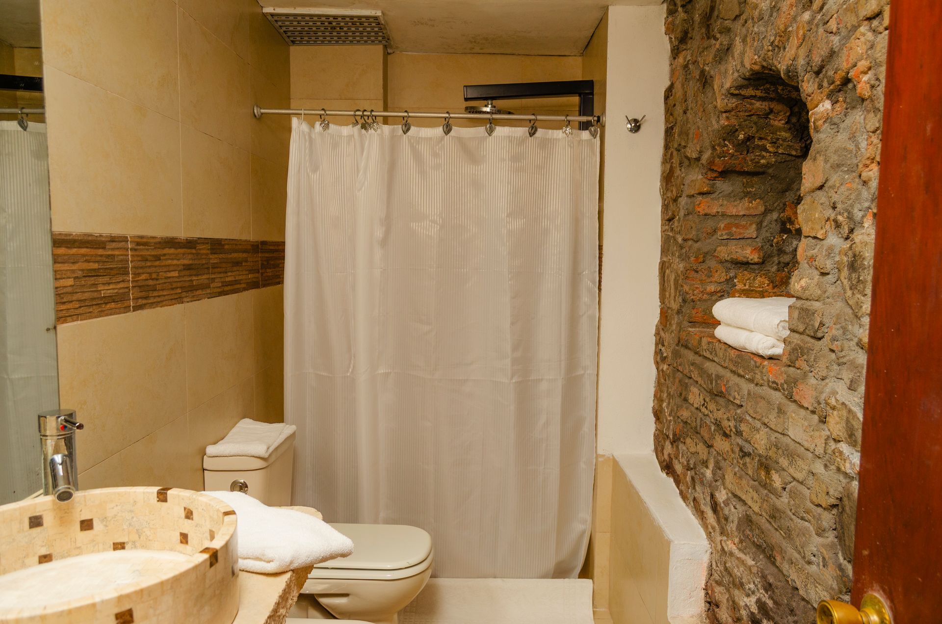 Baño con lavabo sobre encimera, inodoro, ducha con cortina blanca y un nicho en la pared de piedra vista con toallas dobladas.