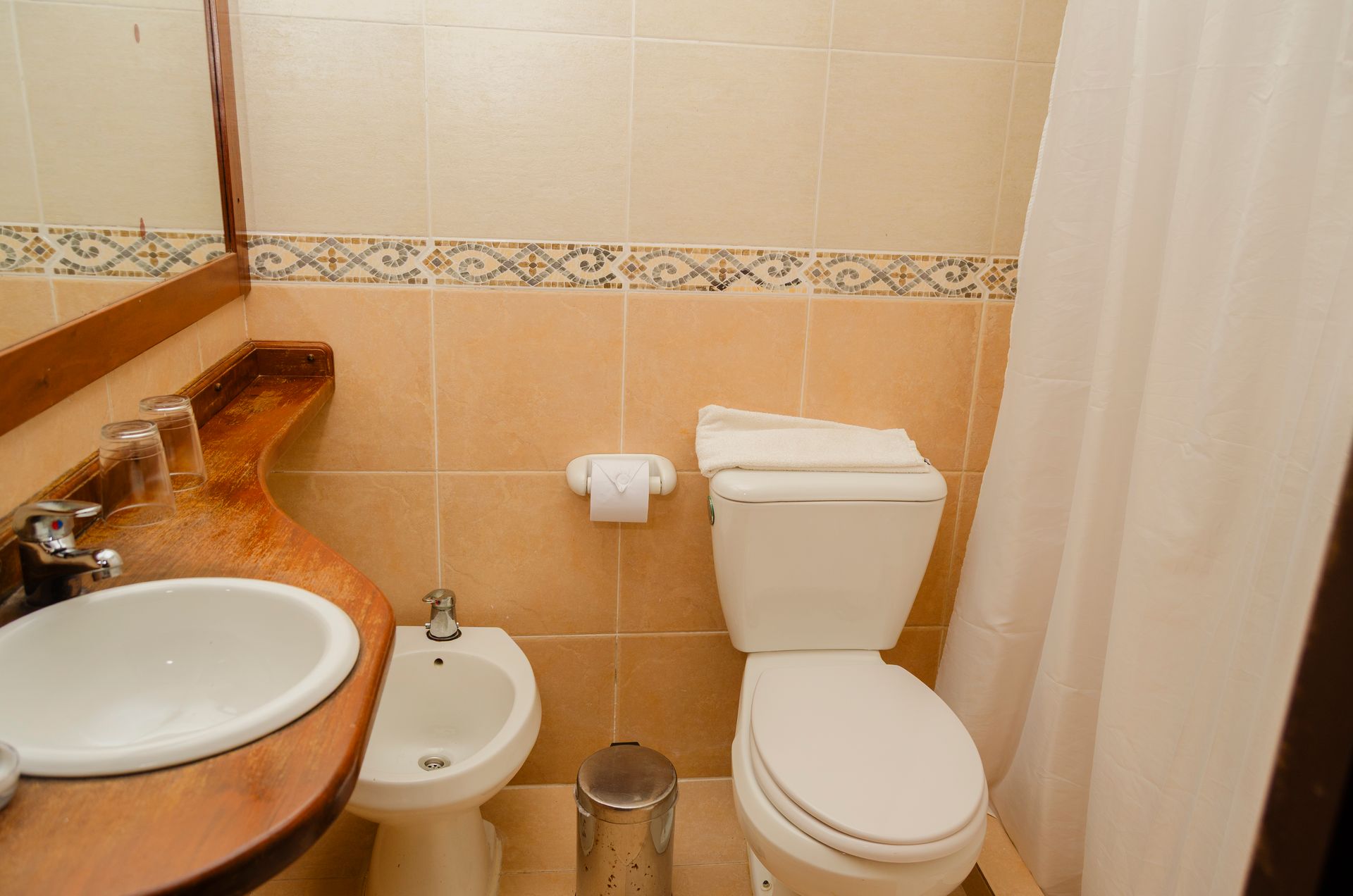 Un pequeño baño de hotel con lavabo blanco, bidé, inodoro y paredes alicatadas en color beige con un borde decorativo.