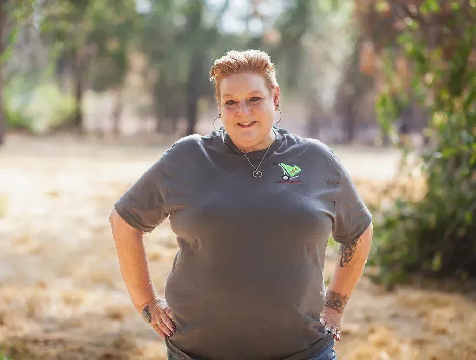 Person standing outdoors on a sunlit dirt path, hands on hips, wearing a dark gray shirt and smiling.