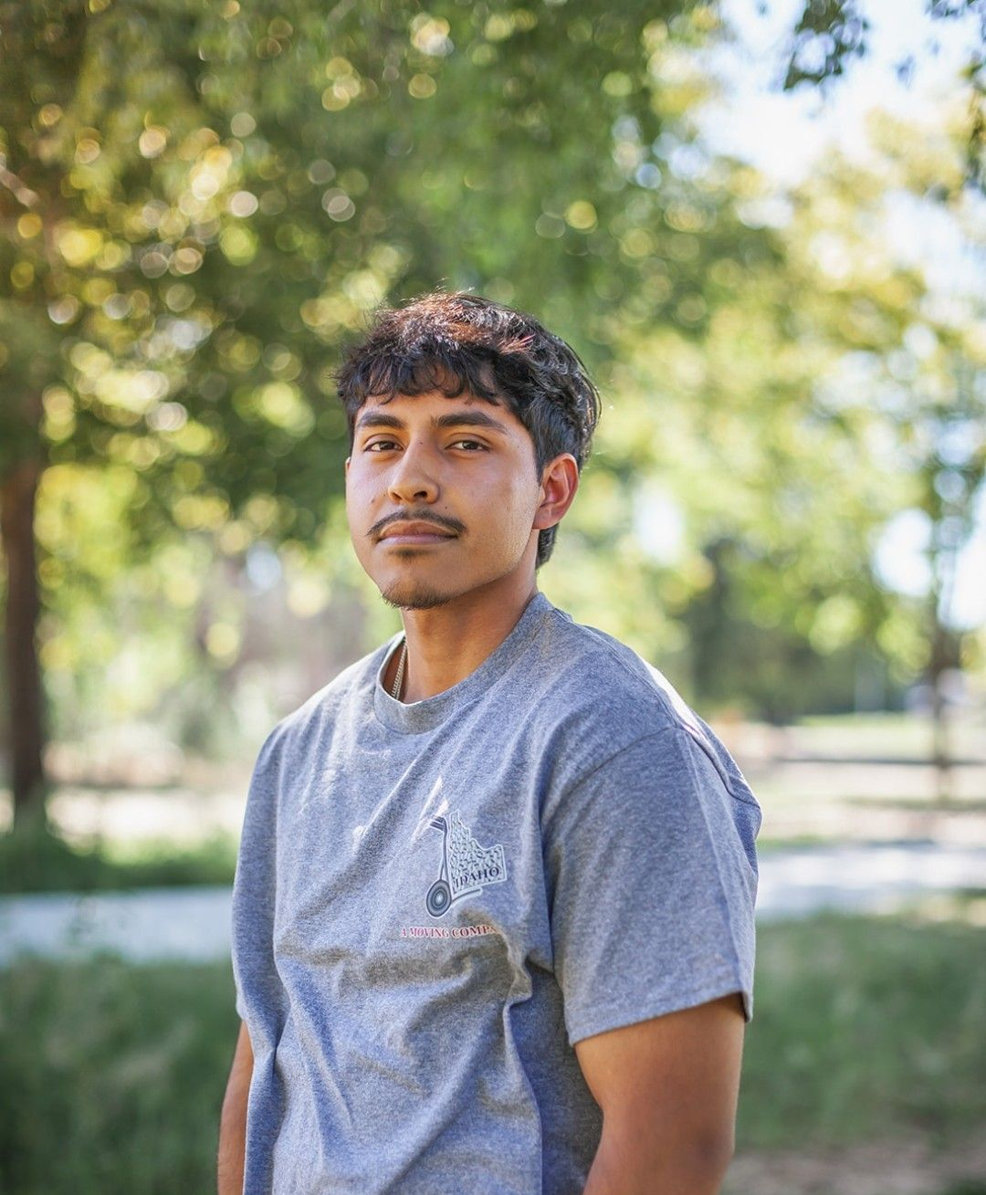 Man standing outdoors in a gray T-shirt, with trees and soft sunlight in the background.