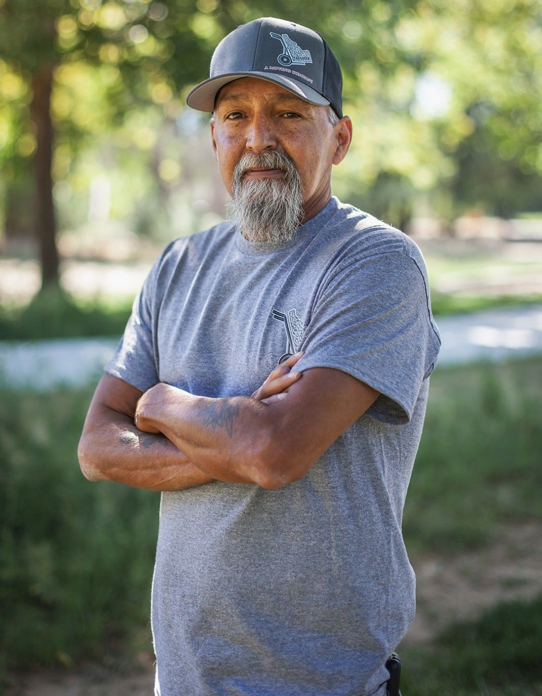 Man in black cap and gray T-shirt with arms crossed, standing outdoors against a blurred green background