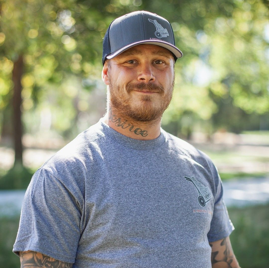 Man in a gray T-shirt and black cap standing outdoors in a park, with tattooed arms.
