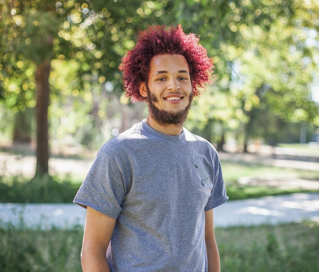 Person standing outdoors in a park, wearing a blue shirt and beige shorts, with curly red hair.