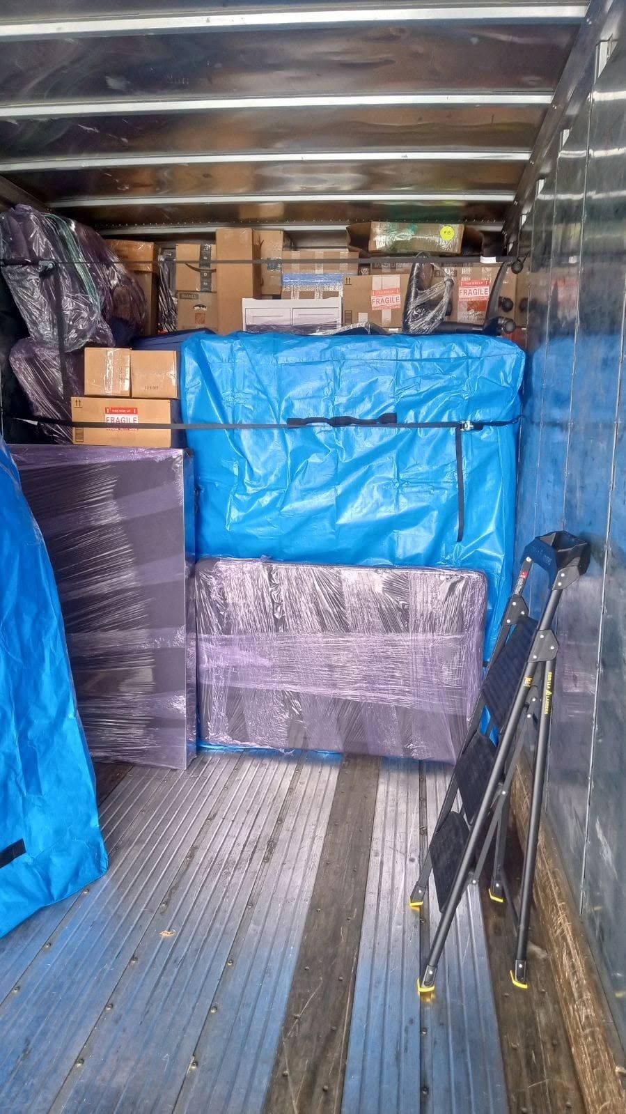 Boxed cargo stacked inside a truck trailer, wrapped in blue and purple plastic.