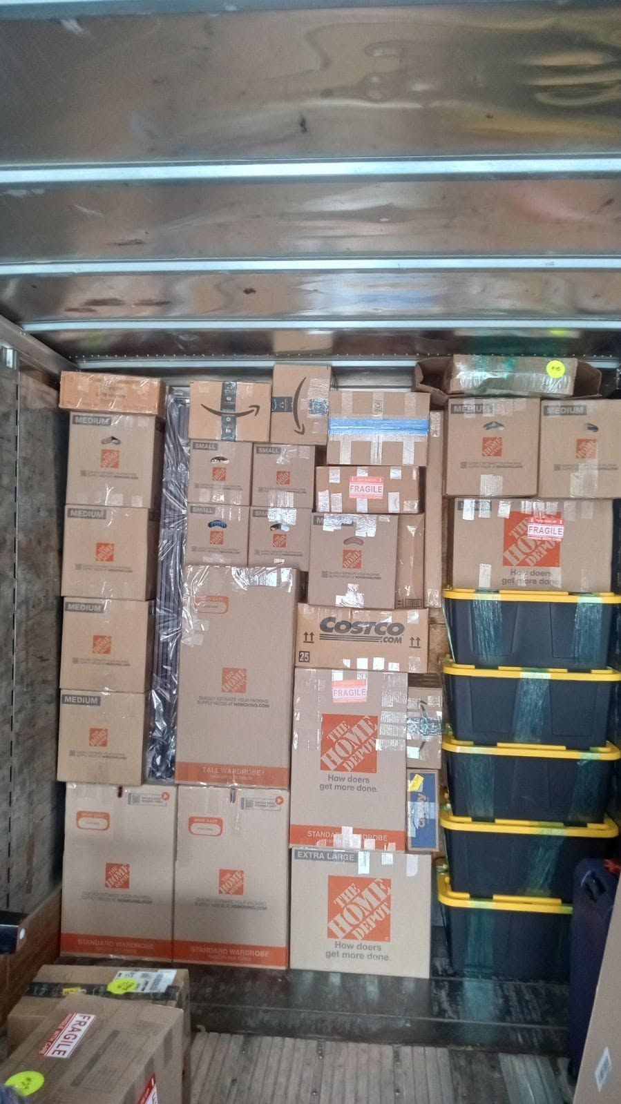 Stacked moving boxes packed inside a truck, with a few blue and yellow storage bins on the right.