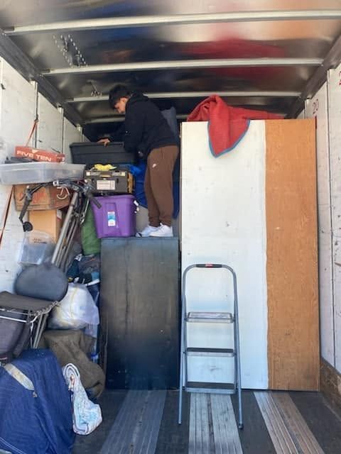 Person standing on stacked items inside a packed moving truck, with a ladder and white cabinet nearby.