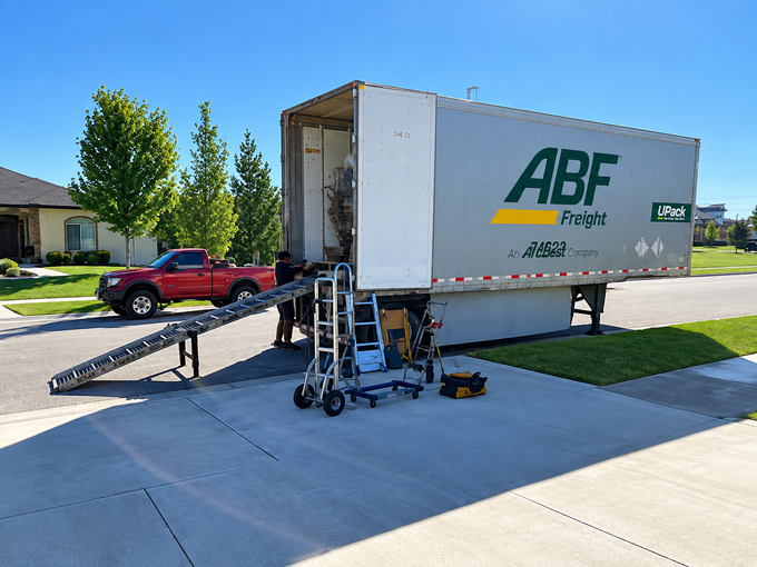 Moving truck with ramp in a driveway, red pickup beside it, under bright blue sky.