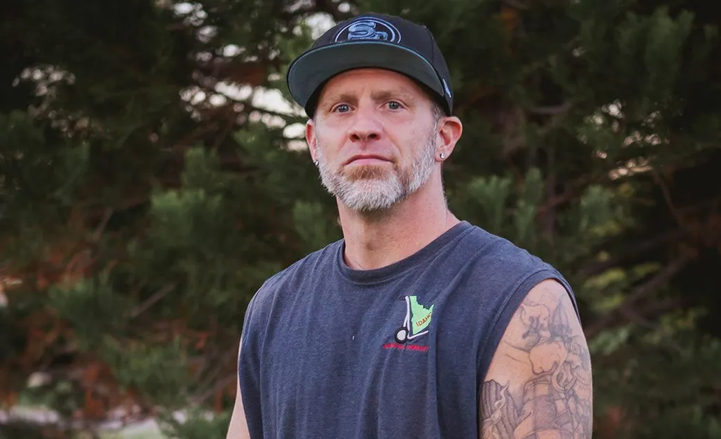 Man in a dark cap and sleeveless shirt standing outdoors in front of trees.