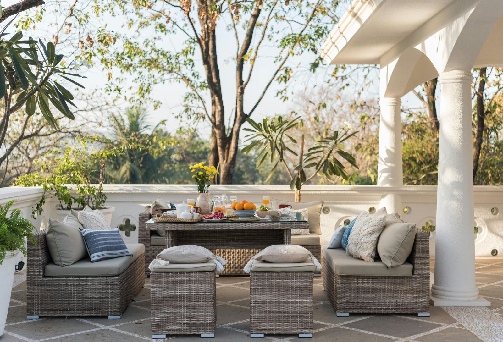 Outdoor patio lounge with wicker sofas, cushions, and a low table under a pergola, surrounded by trees