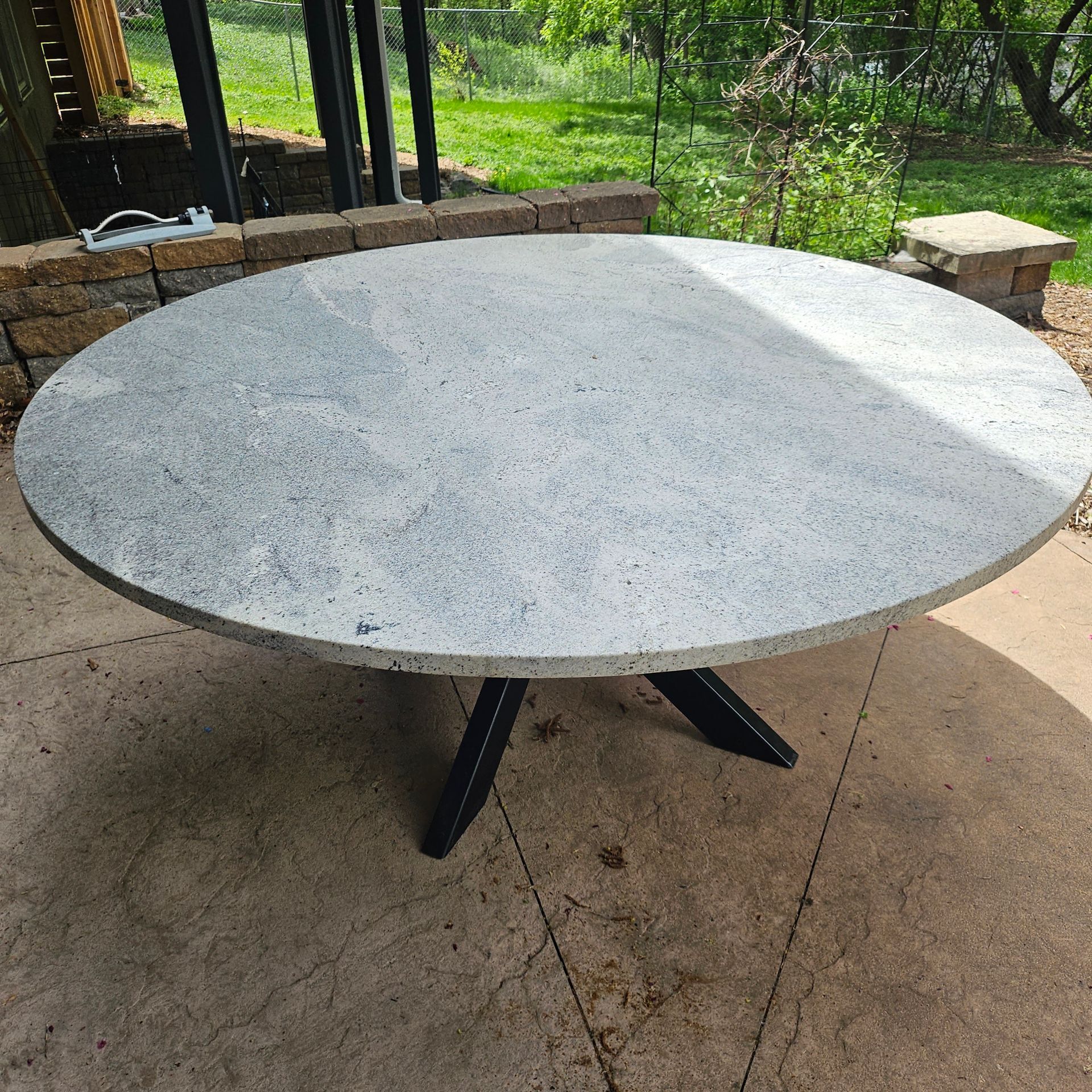 Round speckled stone outdoor table with black pedestal base on a patio, surrounded by greenery