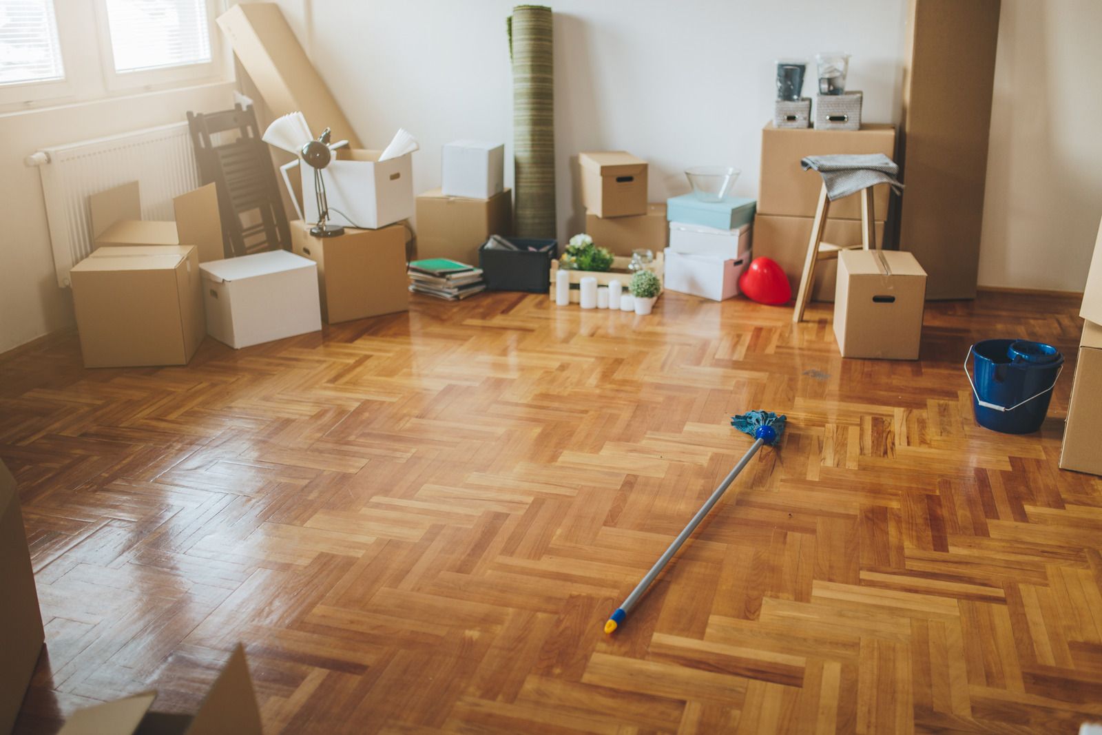 Scattered moving boxes in a sunlit room with wooden floor and a mop on the floor