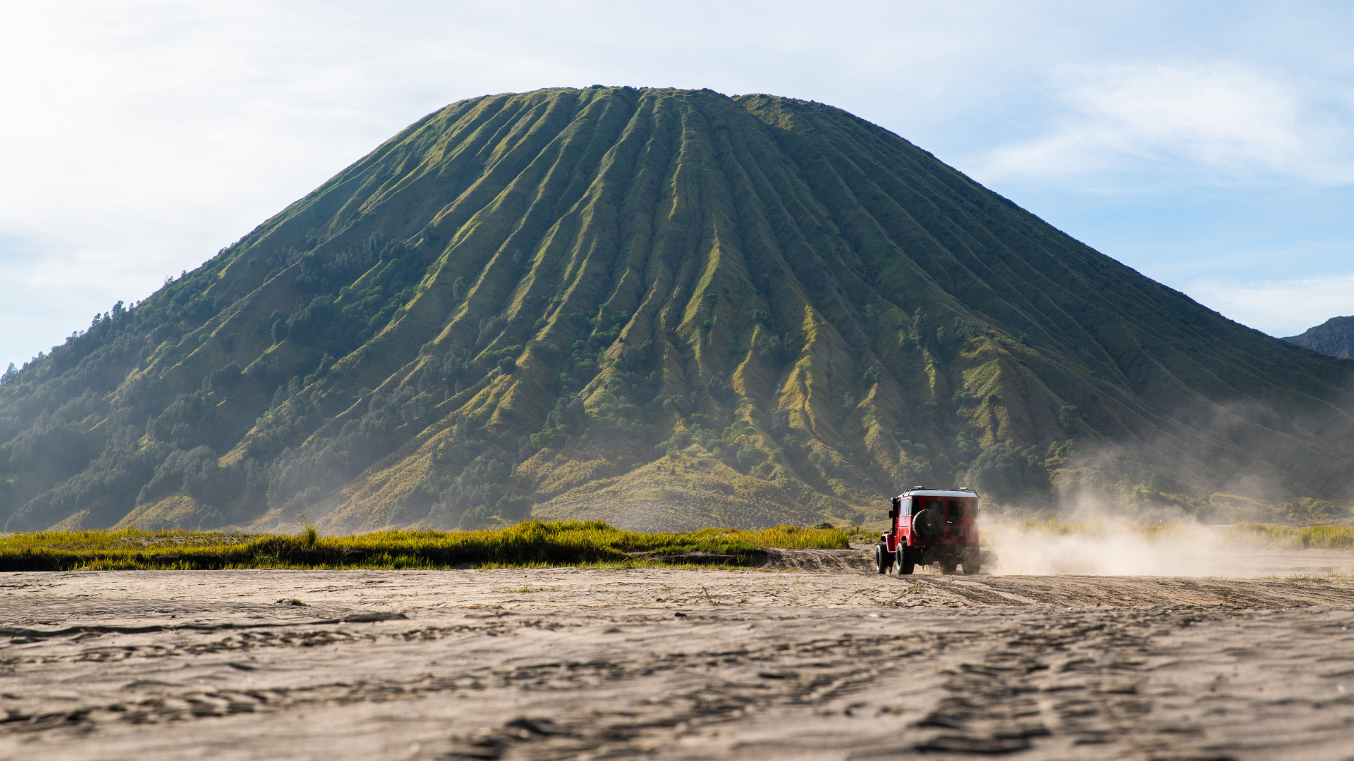 A Jeep kicks up dust as it drives across a sandy plain, Mount Bromo in the background.