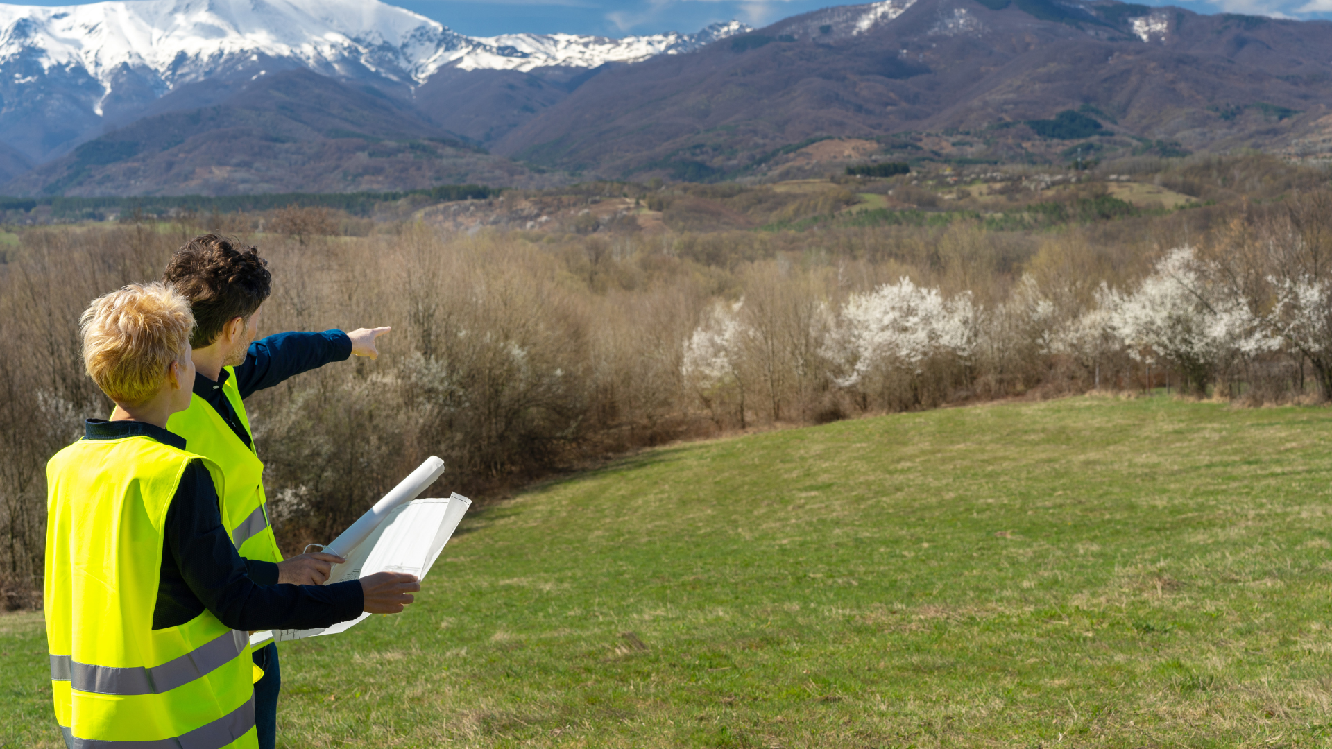 Two people in vests survey land with mountain backdrop.