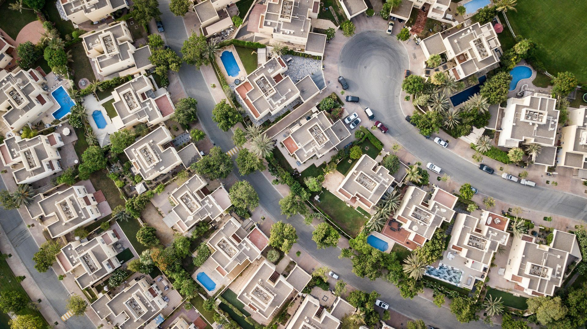Aerial view of a suburban neighborhood with light-colored houses, roads, trees, and swimming pools.