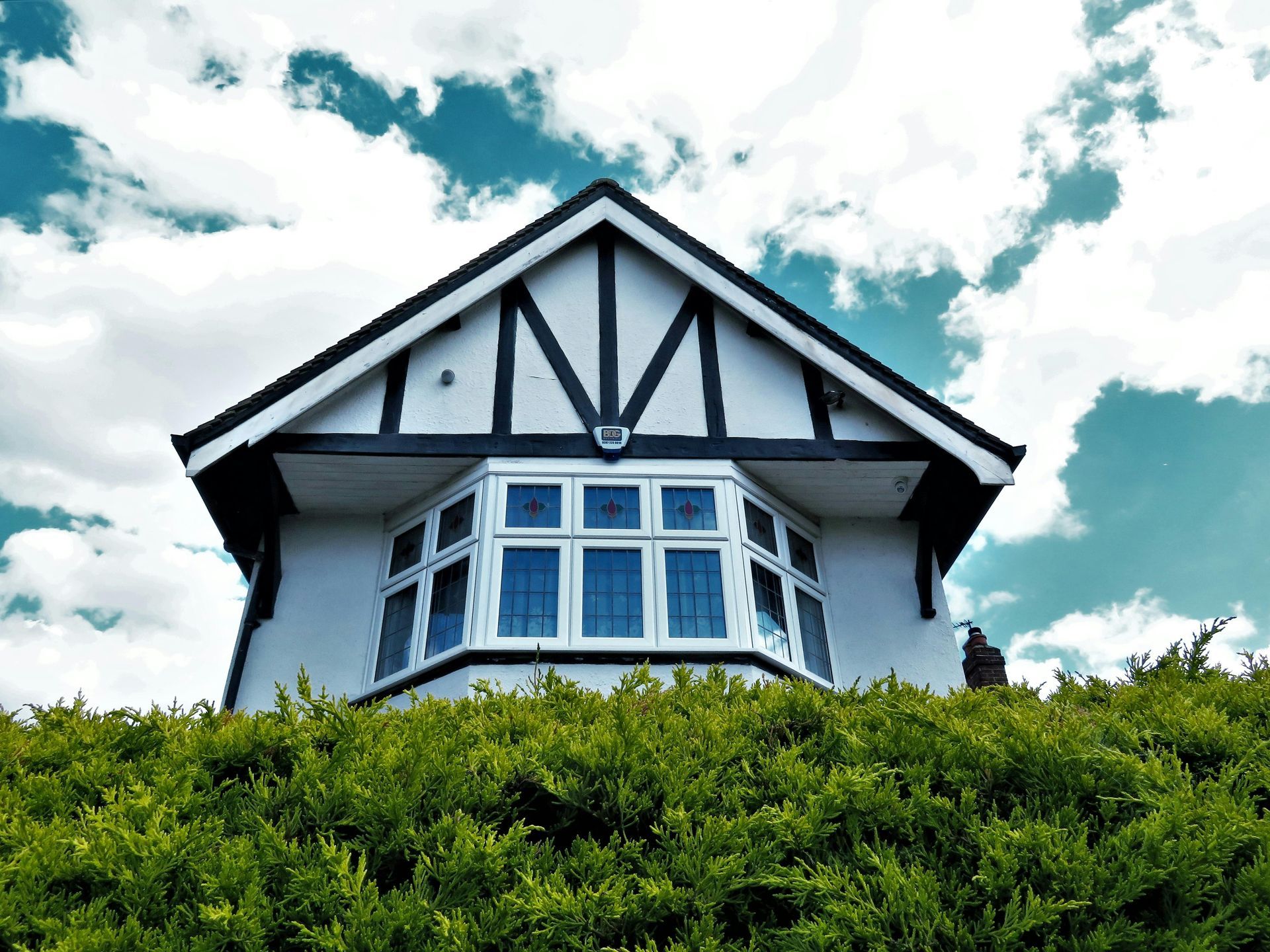 White house with black trim and a bay window, set above green hedge against a cloudy sky.