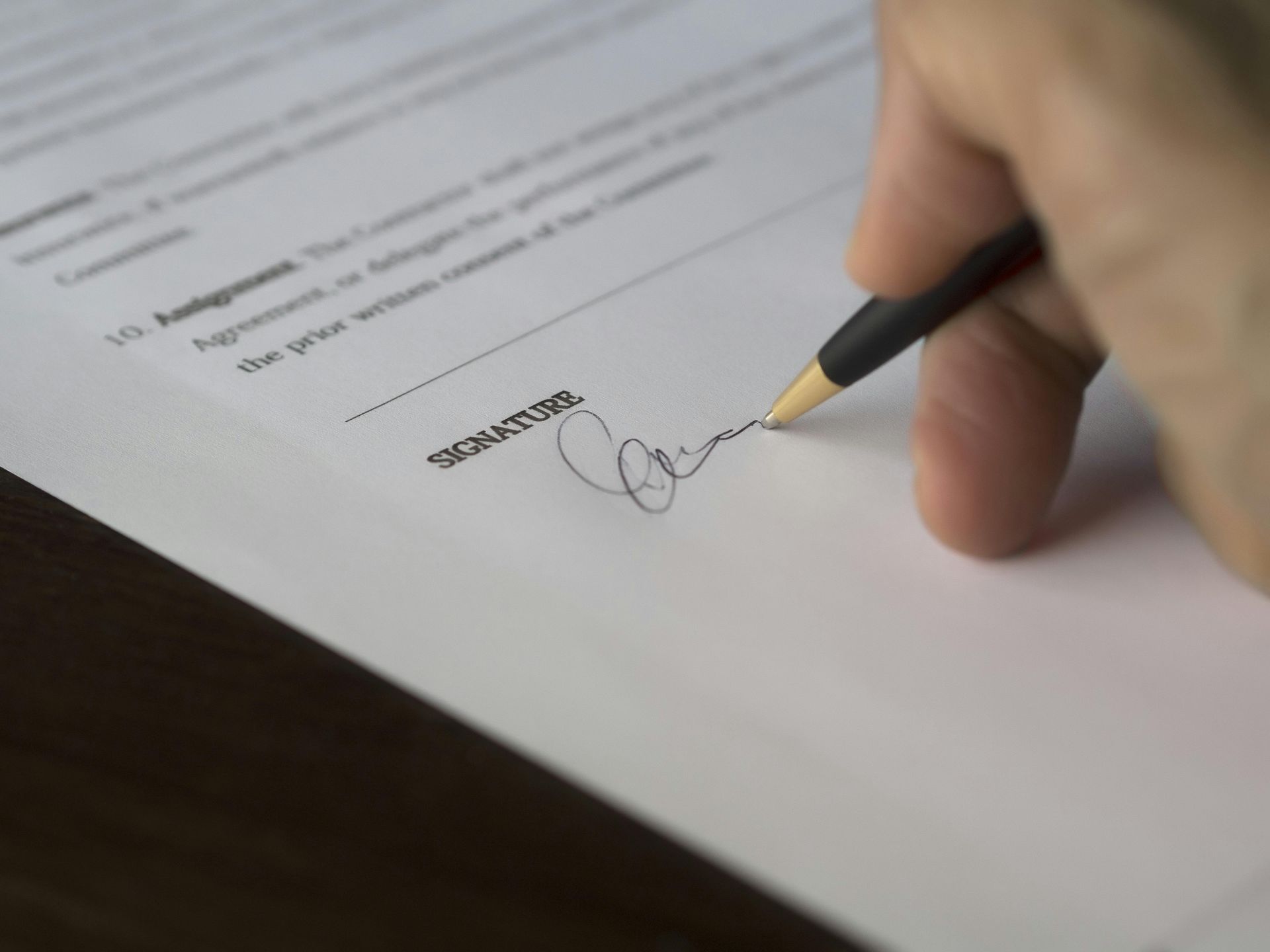 Hand signing a document with a black pen at the signature line.