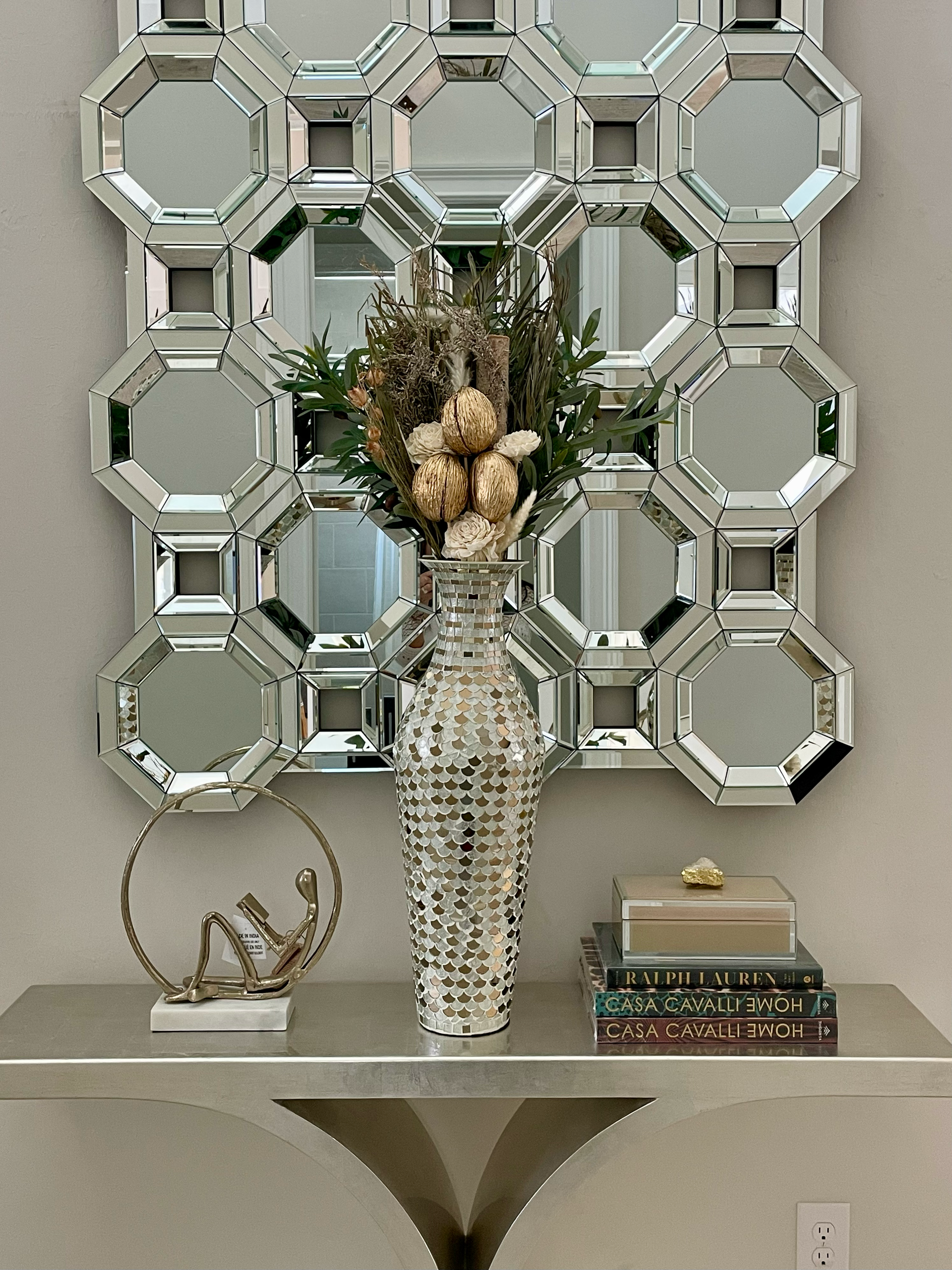 Decorative mirrored wall art above a console table with vase, books, and a small sculpture at CRAFTED 1111 fashion