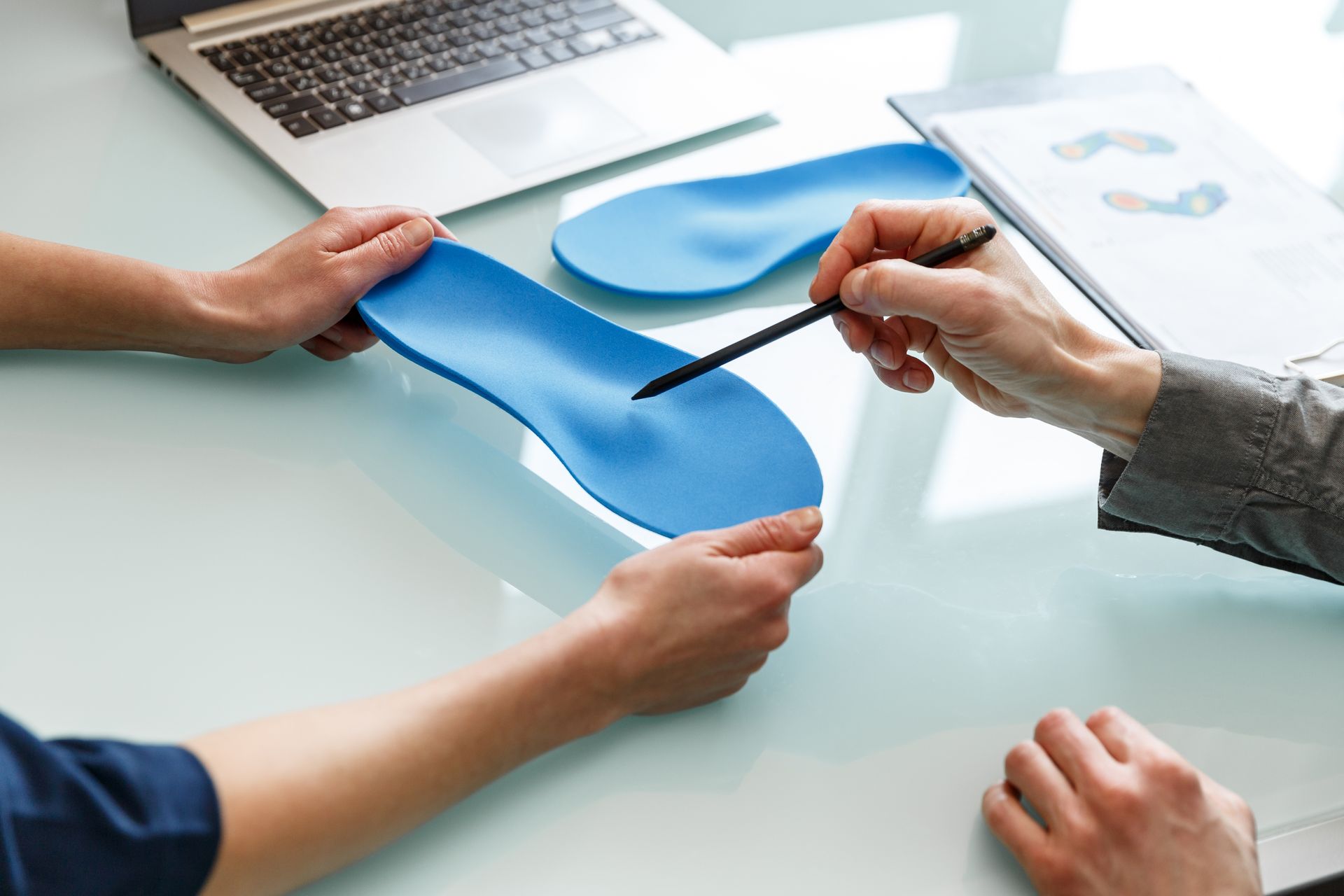 Two people examining blue orthotics on a table, one pointing with a pen. Laptop and foot diagrams present.