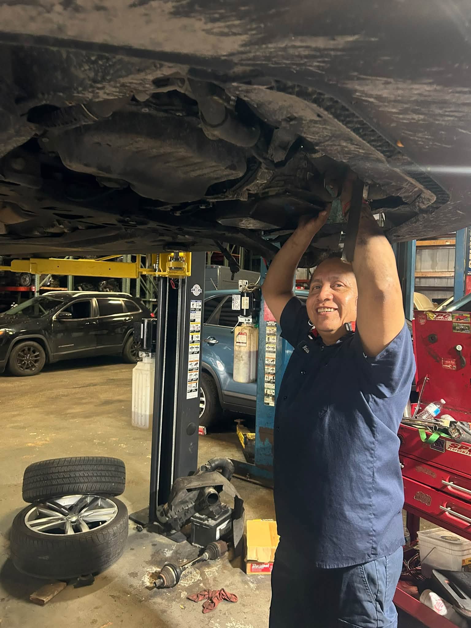 Mechanic smiling under a car on a lift, working in an auto shop with tools and a tire nearby | Auto Logic Complete Auto Repairs