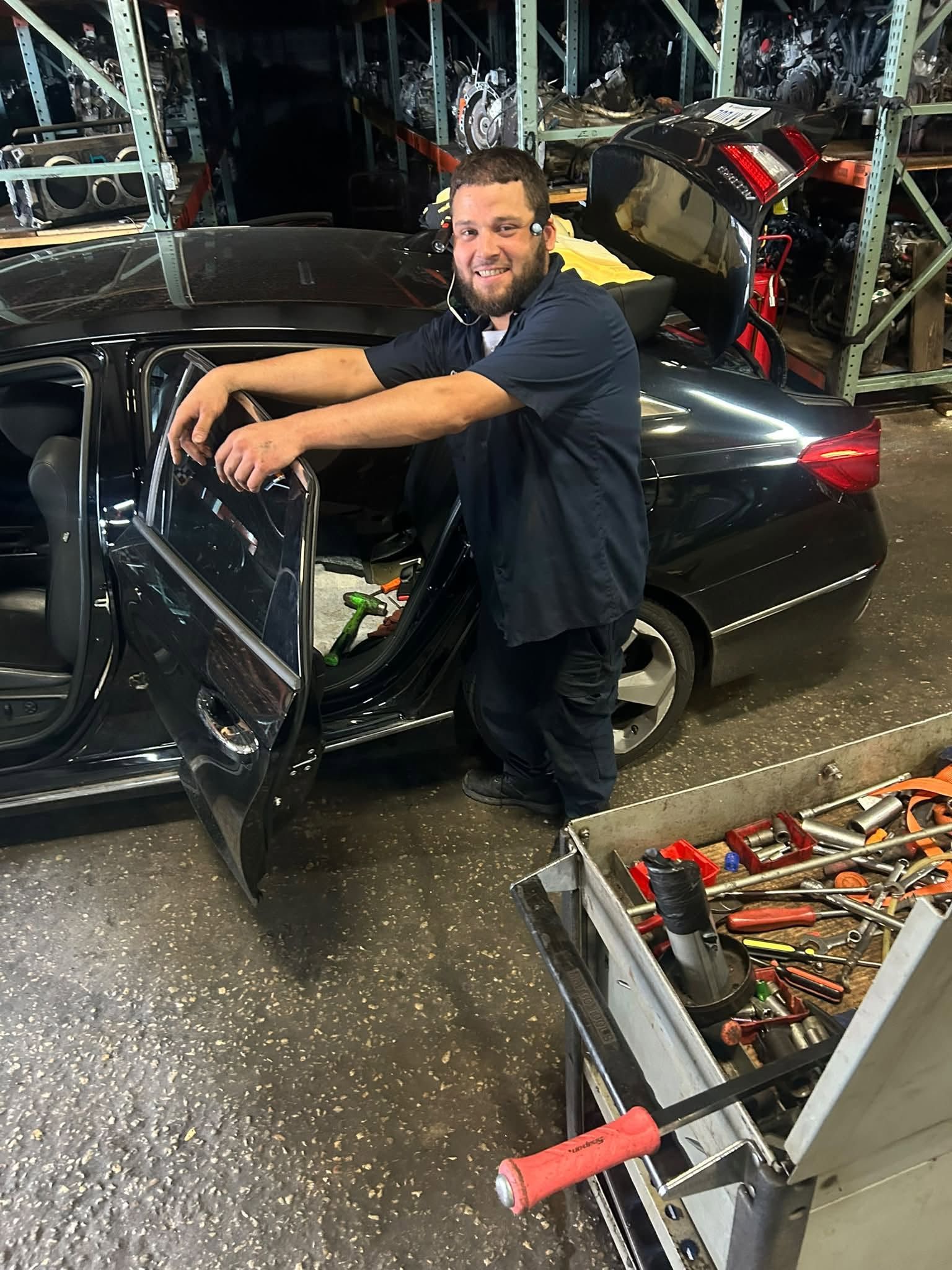 Man smiling beside a black car in a cluttered garage, leaning on the open driver’s door. | Auto Logic Complete Auto Repairs