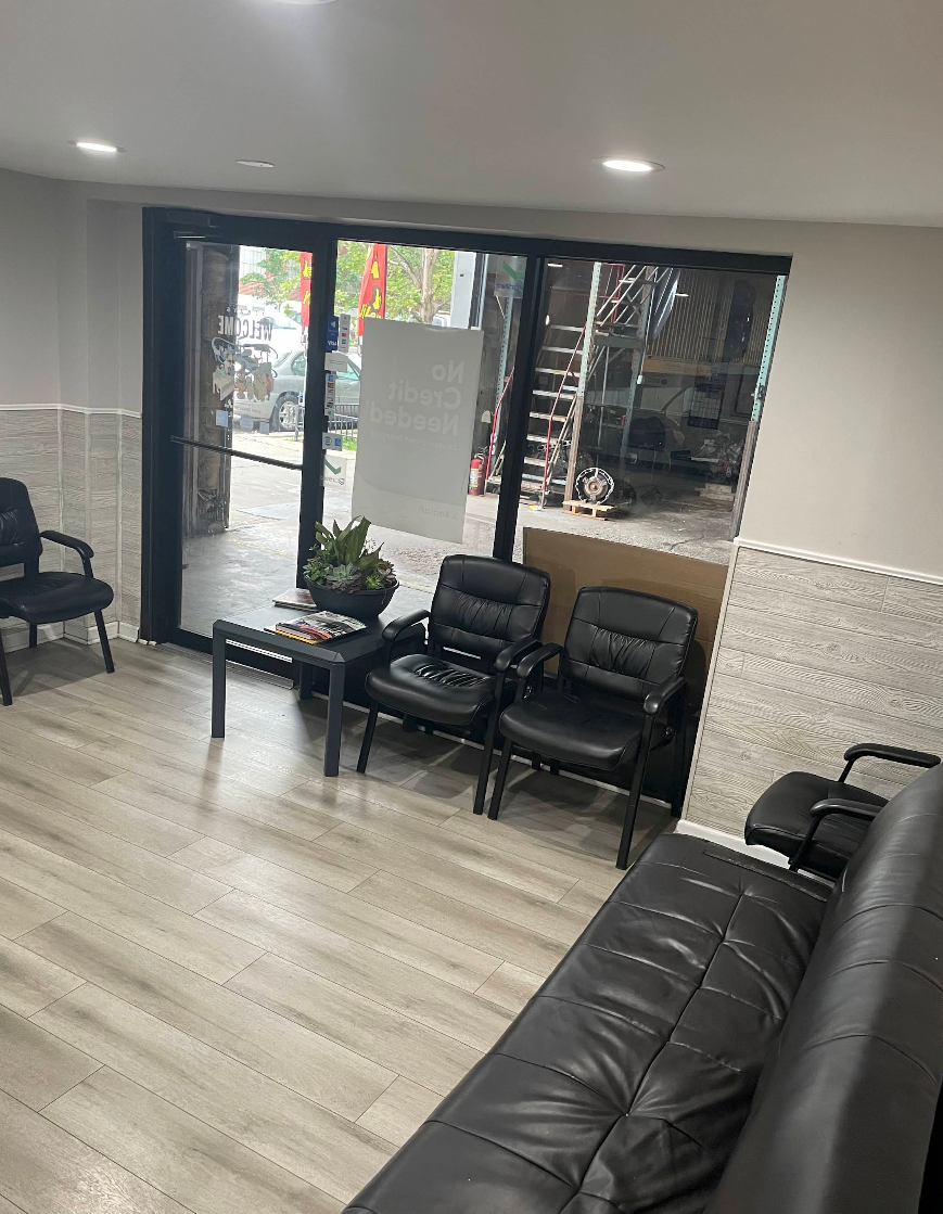 Empty waiting room with black chairs, glass entrance doors, and a dark leather sofa on light wood floors. | Auto Logic Complete Auto Repairs