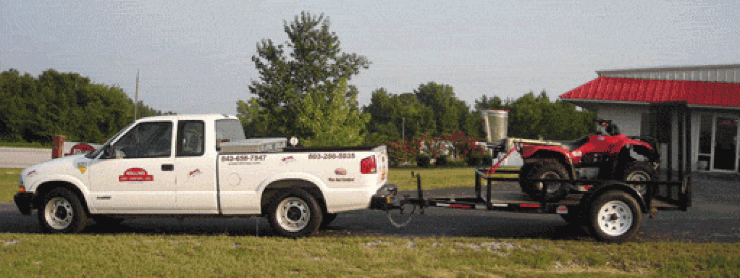 White truck towing a trailer with a red ATV, driving near a building with a red roof.