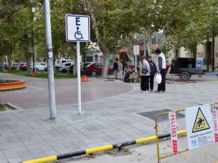 Una señal de aparcamiento para discapacitados se encuentra en una acera cerca de una plaza pública, con barreras de construcción y peatones en las inmediaciones.